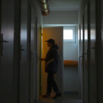 A person silhouetted in a dim hallway opens a door with warm light shining through, evoking a tense atmosphere. The hallway has closed doors on both sides and exposed pipes on the ceiling.