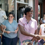 Angie Nixon in a short-sleeve pink shirt greets women while campaigning on a sidewalk.