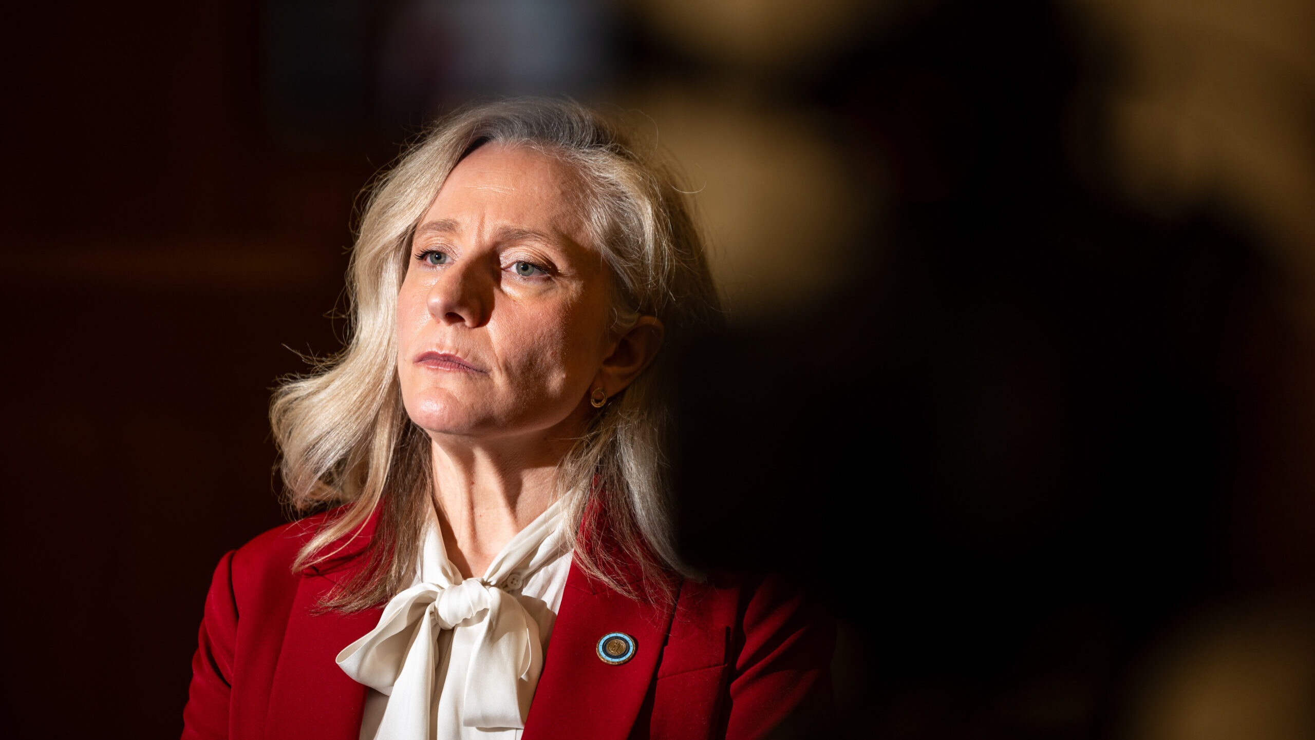 Abigail Spanberger with long gray hair, who wears a red blazer and white blouse, looks slightly to the side. The background is dimly lit with a dark, shadowy area on the right.