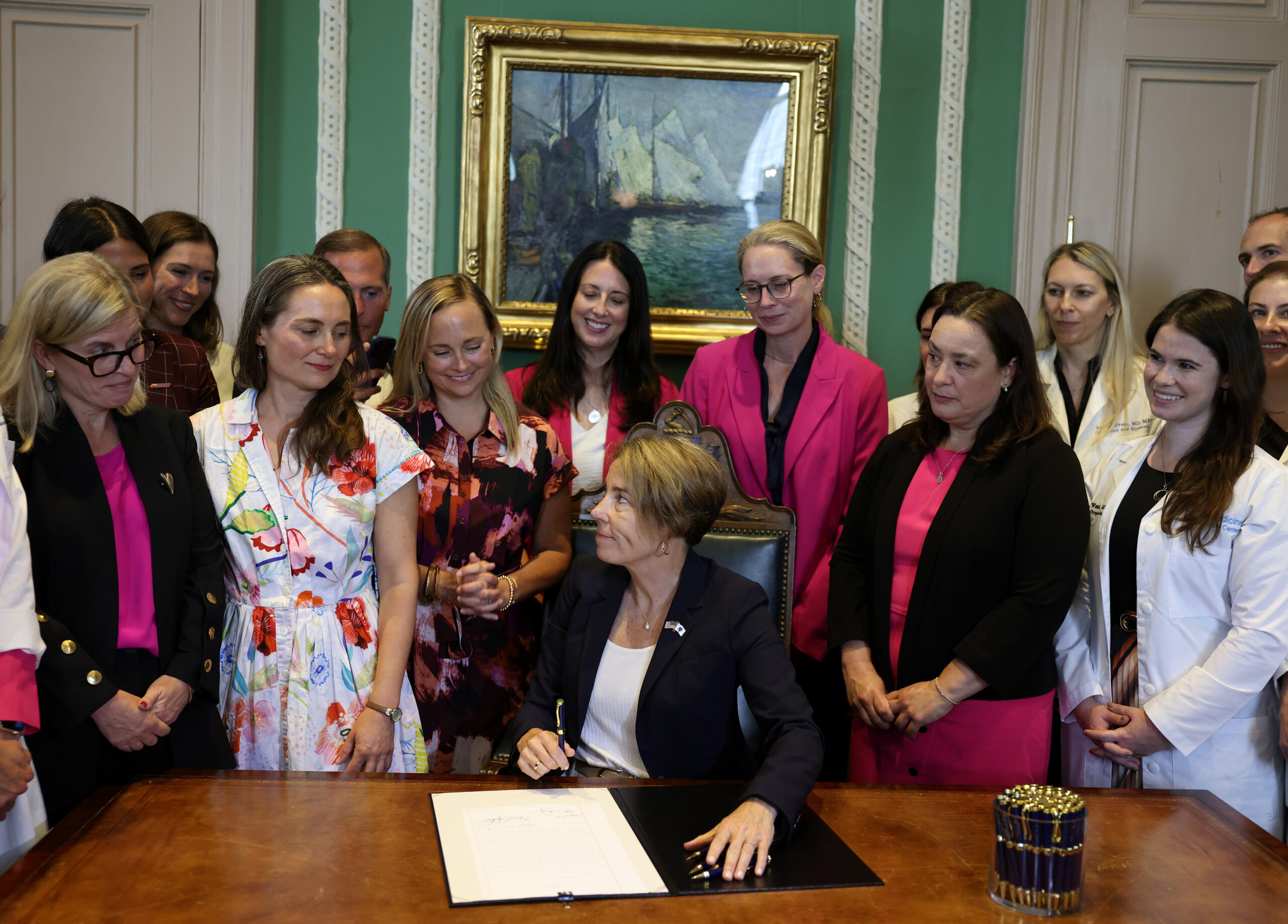 Massachusetts Gov. Maura Healey sits at a desk signing a document related to abortion, surrounded by a group of smiling women and men in professional attire, inside a formal room with a painting in the background.
