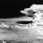 A large mushroom cloud rises over a cityscape following a nuclear explosion, viewed from above with scattered clouds and land visible below.