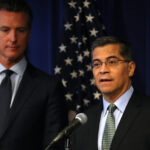 Xavier Becerra and Gavin Newsom in suits stand in front of an American flag; one speaks at a microphone while the other stands beside him, looking on, against a blue curtain backdrop.