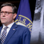 Mike Johnson in a blue suit and striped tie stands in front of U.S. flags, a sign with a black-and-white photo labeled LINE, and what appears to be the entrance to a government house. He appears serious and attentive.