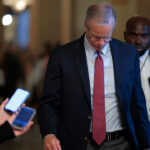 John Thune in a suit and tie looks down while walking indoors, surrounded by people holding smartphones—many of whom appear to be independents covering the event. A security guard walks behind him. The background is softly lit and blurred.