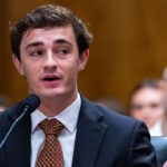 Nick Shirley, a young man in a suit and tie, speaks into a microphone at a public hearing, with people seated and listening in the blurred background.