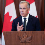 Mark Carney in a suit speaks at a podium with a maple leaf emblem, gesturing with one hand; several large Canadian flags are displayed in the background, emphasizing the official atmosphere of Canada.