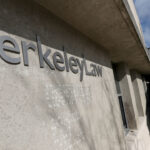 The exterior of Berkeley Law, with large metallic letters on a concrete wall and people walking up adjacent outdoor stairs on a sunny day.