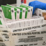 A gloved hand handles a stack of mail-in ballots in green and white envelopes inside a United States Postal Service tray.