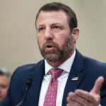 Homeland Security Secretary Markwayne Mullin in a navy suit and pink tie speaks into a microphone at a formal event, gesturing with one hand. He has short brown hair, a beard, and wears an American flag pin on his lapel.