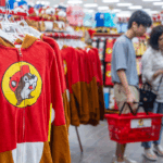 Red and brown animal onesies with a beaver logo hang in a store, while three people with red shopping baskets browse items in the background.