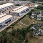 Aerial view showing two large industrial warehouse buildings next to a residential neighborhood with winding roads, single-family homes, trees, and a small pond. Construction equipment and materials are visible near the warehouses, which were recently developed as part of a local tax break initiative encouraging business growth in the area.