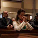 A group of people sits in church pews, heads bowed and hands clasped in prayer, in a softly lit sanctuary.