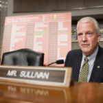 Sen. Dan Sullivan in a suit sits at a desk with a nameplate reading Mr. Sullivan. Behind him is a chart titled Russian & Chinese Incursions Into Alaska in a formal hearing room.