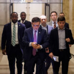 A group of men, including Mike Johnson, in business attire walk down a hallway. The man at the front is looking at his phone, while others follow closely behind. The setting appears to be an official or government building.