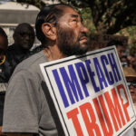A man holds an Impeach Trump sign at a protest, surrounded by people and police officers. Some hold signs, and trees and houses are visible in the background.