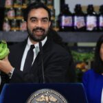 New York City Mayor Zohran Mamdani in a suit at a podium holds up a bunch of green bananas, while a woman in blue stands beside him. Shelves with groceries are visible in the background.
