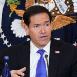 Marco Rubio in a suit speaks at a table with two blue bottles in front of him. Behind him are the U.S. flag and the presidential seal.