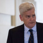 Sen. Bill Cassidy with short gray hair wearing a dark suit, white shirt, and patterned tie stands indoors, looking to his right with a neutral expression.