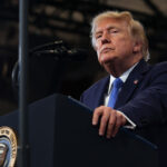 President Donald J. Trump wears a dark suit and blue tie stands at a podium with a presidential seal, speaking into a microphone in a well-lit room.