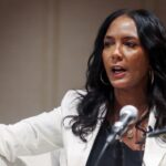 A woman with long dark hair and a white blazer speaks passionately into a microphone, gesturing with one hand while seated on a light-colored chair.