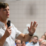 Rob Sand in a striped shirt holds a microphone and speaks, gesturing with his hand, in front of a seated audience in a room with tiled walls.