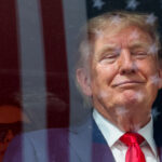 President Trump in a suit and red tie smiles while standing in front of a window with the reflection of the American flag. Another person with sunglasses is partially visible in the background.