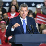 Man in a blue suit speaking at a podium with microphones, addressing a crowd in front of a patriotic backdrop featuring stars and stripes. Audience members are seated and appear to be listening.