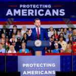 Vice President JD Vance in a suit speaks at a podium labeled PROTECTING AMERICANS with a crowd seated behind him and a large sign displaying the same message.