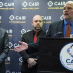 Three men stand at a CAIR podium with microphones. Behind them is a CAIR banner and a U.S. flag. One man is speaking, while another gestures with his hand.