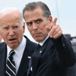 Two men in suits stand outdoors. One is looking forward with his mouth slightly open, while the other gestures and points ahead. A third man in sunglasses stands behind them. A vehicle is visible in the background.