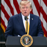 A man in a suit speaks at a podium with the U.S. presidential seal, with several large American flags in the background.