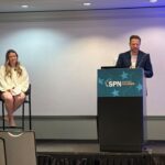 Camille Walsh, director of media relations at State Policy Network, sits on a tall chair, and Corey Burres, senior director of communication strategies at State Policy Network, stands at a podium labeled SPN Orlando in a conference room, with empty chairs in the foreground.