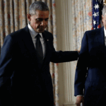 Then-President Barack Obama and then-Attorney General Eric Holder stand together indoors, one with his hand on the others back. U.S. flags and curtains are in the background. Another person claps in the background on Sept. 25, 2014.