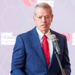 Nebraska Gov. Jim Pillen in a blue suit and red tie speaks into a microphone at a podium, with a blurred backdrop featuring the UNMC Nebraska Medicine logo.