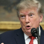 President Donald Trump in a dark suit speaks into a microphone, gesturing with his hand. He has light hair and is standing in front of a blurred painting or background.