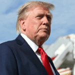 President Donald Trump in a suit and red tie stands outdoors with an airplane staircase in the background and a blue sky overhead.