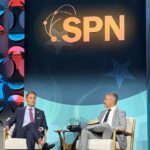 Former Virginia Attorney General Jason Miyares sits on stage alongside State Policy Network's Tony Woodlief under a large SPN sign, engaged in conversation. The backdrop features a blue and red geometric pattern and spotlights. A small table with water glasses is between them.