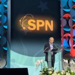 Speaker Yuval Levin stands at a podium on a decorated stage with the letters SPN prominently displayed behind him. Flowers are arranged in front of the podium, and the background features a geometric pattern with blue and red lights.