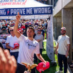 Charlie Kirk stands among a crowd tossing red hats, with people reaching for them. Behind him is a tent with PROVE ME WRONG printed on it. Many in the crowd wear red hats and sunglasses, and the scene is outdoors on a sunny day.