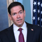 Marco Rubio in a suit and red patterned tie speaks in front of a U.S. flag and a painting, with an American flag pin on his lapel.