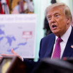 A man in a suit speaks while seated at a desk, gesturing with his hands. A map of the Great Lakes is visible in the blurred background, along with flags and ornate decor.