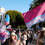 People march outdoors, some holding a large trans pride flag with a Soviet symbol. A person wears butterfly wings, and buildings, palm trees, and more flags are visible in the background. It is sunny and crowded.