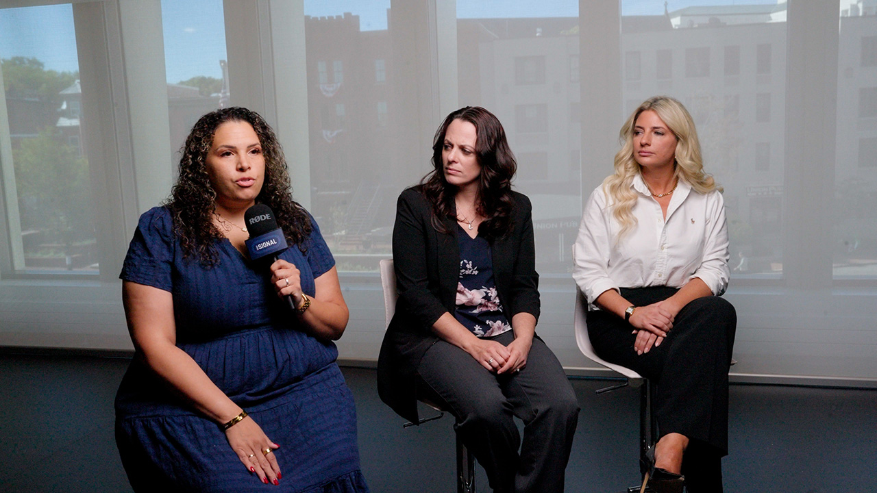 Three women sit indoors on stools; the woman on the left holds a microphone, while the other two listen intently. All are dressed in business casual attire, engaged in a thoughtful discussion about abortion. Large windows with a city view are in the background.
