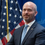 A conservative man in a suit and tie stands in front of an American flag and a blue backdrop, speaking at a podium.