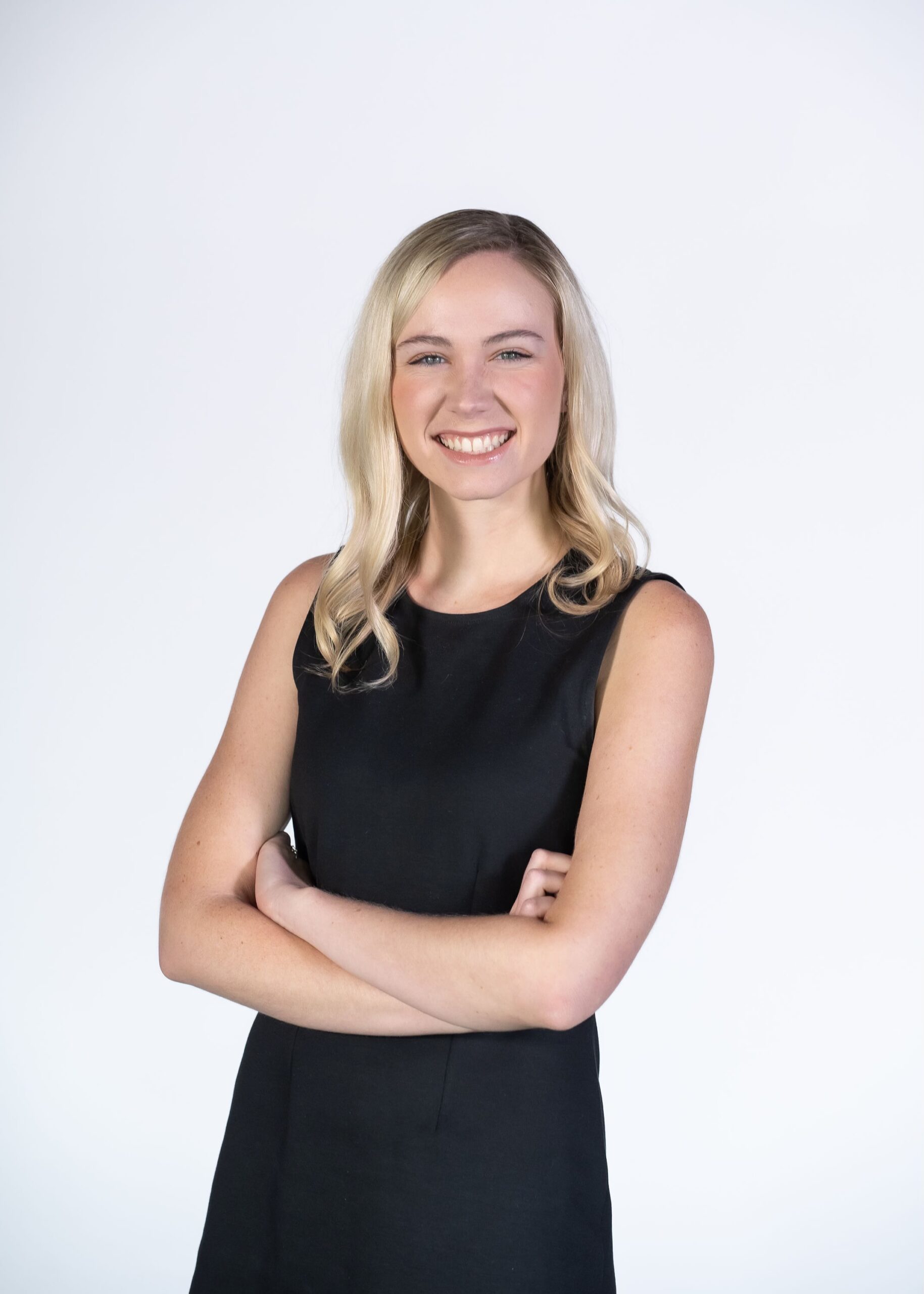 A woman with long blonde hair, wearing a sleeveless black dress, stands against a plain white background, smiling with her arms crossed—confident and ready for her Daily Signal internship.