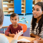Parental rights concept. Mother speaking with teacher in school. A woman and a young boy sit across from another woman at a table, engaged in conversation in a classroom setting. The boy looks curious while the women appear to be discussing something important.