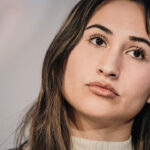 Detransitioner Chloe Cole reveals the dangers of "gender-affirming care" and pediatric gender interventions. Pictured: Cole, a young woman with long brown hair and a neutral expression looks slightly upward. She is wearing a light-colored top and is photographed against a blurred background.