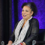 Pictured: Professor Melina Abdullah, a woman with curly hair, sits on a chair, smiling. She wears a black outfit with a white patterned scarf. Behind her is a purple-lit curtain. A water bottle and green glass bottle are on a table nearby.