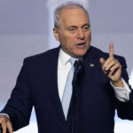 A man in a dark suit and light blue tie speaks at a podium, gesturing with his right hand raised and index finger pointing up. A blurred background shows large white stars.