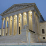 The U.S. Supreme Court building at dusk, illuminated by lights, with tall columns, marble steps, statues, and part of a fountain visible in the foreground.